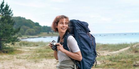 Eine Frau mit Wanderrucksack steht am Seeufer und hat eine Fotokamera in der Hand