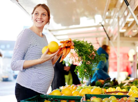 Schwangere Person mit Gemüse in der Hand vor einem Gemüsestand.