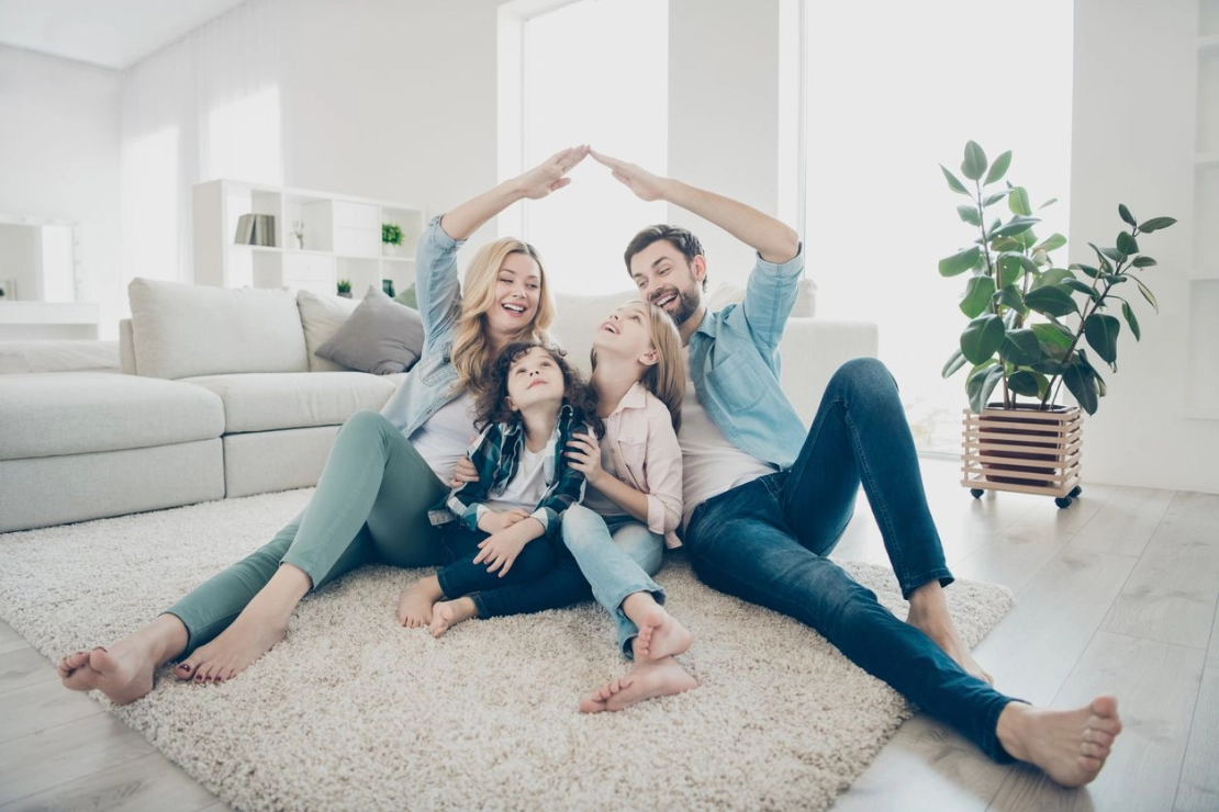 Familie sitzt auf Teppich im Wohnzimmer, bildet Dach mit Händen über Köpfen, modernes helles Interieur mit Sofa und Pflanze