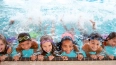 Mehrere Kinder in bunten Schwimmwesten halten sich am Beckenrand eines Pools fest und lächeln in die Kamera.