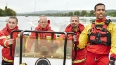 Vier Personen in roter und gelber Rettungsausrüstung halten eine Sonarbildschirm an einem Fluss, dahinter grüne Landschaft und Brücke.