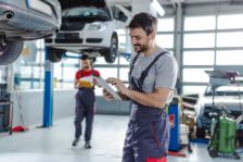 In einer KFZ-Werkstatt stehen stehen Autos auf einer Hebebühne. Zwei Kfz-Mechaniker arbeiten in der Werkstatt,  einer der Kfz-Mechaniker tippt etwas in sein Tablet .