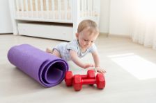 Ein Kleinkind krabbelt neben einer Yogamatte auf dem Fußboden und spielt mit einem Paar roter Hanteln