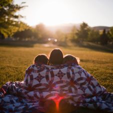 Kinder sitzen auf einer Wiese in eine Decke gehüllt und schauen in den Sonnenuntergang
