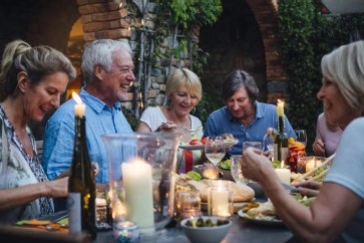 Fünf Personen sitzen beim Abendessen an Tisch mit Kerzen, Flaschen und Speisen unter Backsteingewölbe mit Efeu.