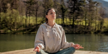 Frau im Trainingsoutfit sitzt am See und praktiziert Yoga.