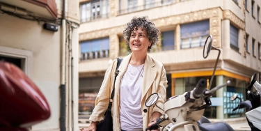 Eine Frau bei ihrem geparkten Moped symbolisiert den Schutz vor Diebstahl durch eine zusätzliche Teilkaskoversicherung.