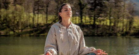 Frau im Trainingsoutfit sitzt am See und praktiziert Yoga.
