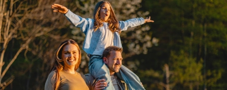 Familie wandert durch den Wald, der Vater trägt die Tochter auf den Schultern.