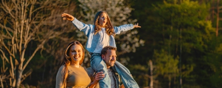 Familie wandert durch den Wald, der Vater trägt die Tochter auf den Schultern.