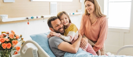 Vater liegt in einem hellen freundlichen Krankenhauszimmer im Bett und wird von seiner Frau und Tochter besucht.