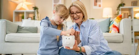 Großmutter und Enkelin werfen Münz-Geld in ein Sparschwein als Symbol für das Sparen für Kinder.