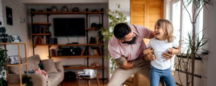 Vater bringt und junger Tochter im Wohnzimmer das Skateboardfahren bei.