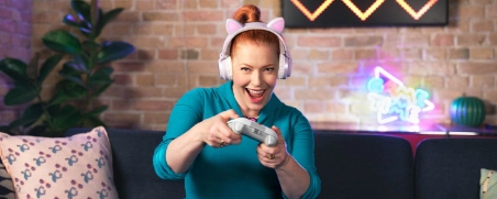 Lachende Frau in blauem Kleid sitzt auf Sofa, macht Gaming-Gesten, Ziegelwand mit Neon-Herzen und Pflanze im Hintergrund