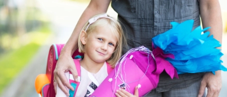 Ein Mädchen hält eine Schultüte zum ersten Schultag und lächelt in die Kamera. Im Hintergrund der Vater, der den Arm um sie gelegt hat.