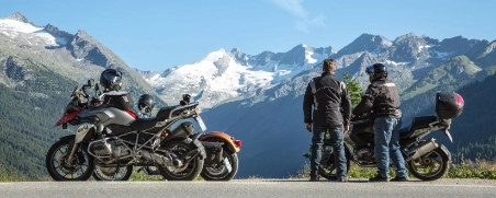 2 Freunde sind mit dem Motorrad im Ausland unterwegs. Sie haben angehalten und blicken auf die Berge