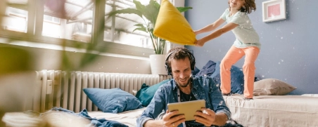 Vater hört Music auf dem Tablet und Kind tobt auf dem Sofa und spielt Kissenschlacht.