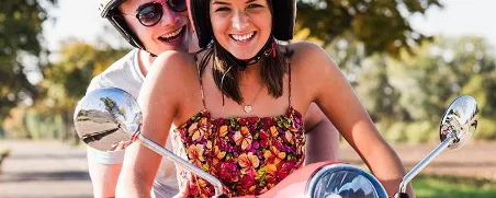 Junge Frau sitzt auf Ihrer Vespa mit einem Freund als Sozius und denkt mit Freude, dass Sie mit dem Moped gut versichert ist.