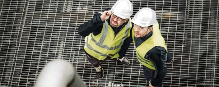 Zwei Bauarbeiter in Schutzwesten und Helmen blicken von oben auf Metallgitter herab und lächeln.