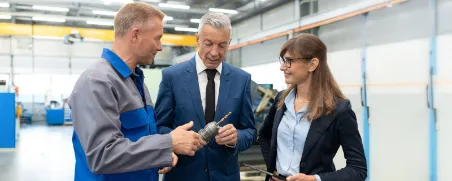 Eine Unternehmerin und ein Unternehmer stehen in einer Werkhalle und besprechen ein Projekt im Vordergrund laeuft ein Mitarbeiter an der Gruppe vorbei