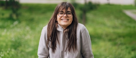 Junge Frau mit langen dunklen Haaren und Brille sitzt im Park auf einer Bank und lächelt in die Kamera