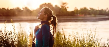 Frau mit braunen Haaren in Sportbekleidung läuft am Ufer eines Sees in Abendstimmung und streckt ihr Gesicht in Richtung Sonne.