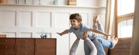 Vater liegt auf dem Rücken und hebt seinen Sohn spielerisch in die Luft im hellen Wohnzimmer