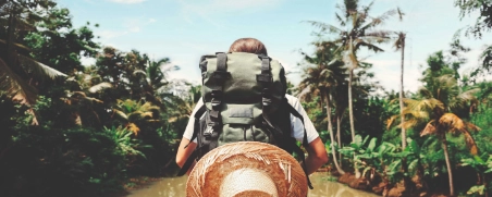 Person mit Rucksack und Strohhut sitzt in Boot auf Fluss, tropische Vegetation und Palmen im Hintergrund