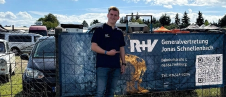 Mann steht lächelnd vor blauem Werbebanner der R+V Generalvertretung Jonas Schnellenbach auf einer Veranstaltung im Freien.