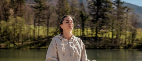 Frau im Trainingsoutfit sitzt am See und praktiziert Yoga.
