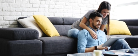Mann sitzt am Laptop auf grauem Sofa, Frau lehnt dahinter, gelbe Kissen, Wohnzimmer mit Ziegelmauer