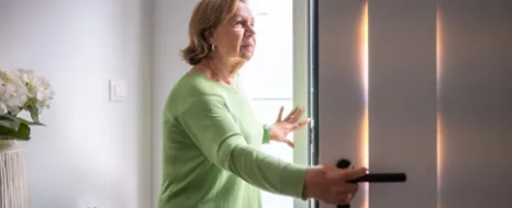 Aeltere Frau in gruenem Pullover die Haustuer in hellem Innenbereich oeffnet