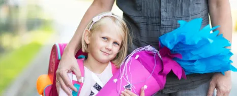 Ein Mädchen hält eine Schultüte zum ersten Schultag und lächelt in die Kamera. Im Hintergrund der Vater, der den Arm um sie gelegt hat.