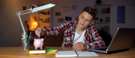 Ein Teenager sitzt an seinem Schreibtisch und tut Geld in sein Sparschwein.