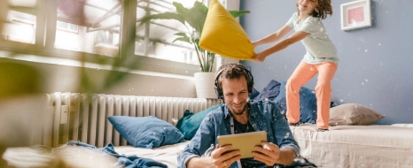 Vater hört Music auf dem Tablet und Kind tobt auf dem Sofa und spielt Kissenschlacht.