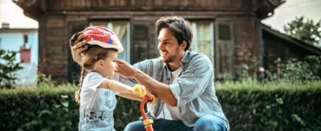 Ein Vater setzt seiner Tochter auf dem Fahrrad einen Helm auf als Symbol für die Kindervorsorge durch die Eltern. 