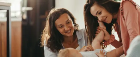 Eine junge Mutter betrachtet liebevoll gemeinsam mit einer Freundin ihr Baby. Die Freundin ist die Patentante des Babys. Sie denken über das Sparen für Patenkinder nach.