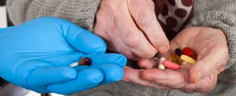 Senior mit Tabletten in der Hand nimmt noch mehr Tabletten aus der Hand einer Pflegeperson mit Handschuhen an.