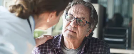 Weibliche Pflegefachperson beugt sich zu pflegebeduerftigem aelterem Mann Hand auf die Schulter legend