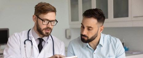 Arzt in Kittel und Brille bespricht mit Patient Befunde auf Tablet in Klinik