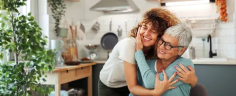 Tochter umarmt sitzende Mutter liebevoll am Esstisch.