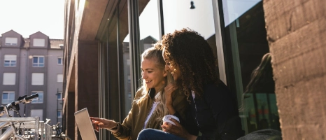 Zwei Frauen sitzen am Fenster und arbeiten gemeinsam am Laptop, Stadtkulisse im Hintergrund
