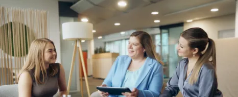 Drei Kolleginnen sitzen zusammen und unterhalten sich mit einem Tablet in der Hand.