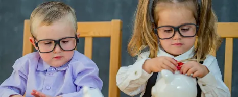 Zwei Kinder in Erwachsenenkleidung sitzen am Tisch; das Mädchen steckt Münzen in ein Sparschwein, während der Junge einen Taschenrechner hält und Münzstapel vor ihnen liegen.