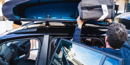 Familienvater mit grauem Haar, der einen Reisetasche in eine Dachbox auf seinem Auto packt.