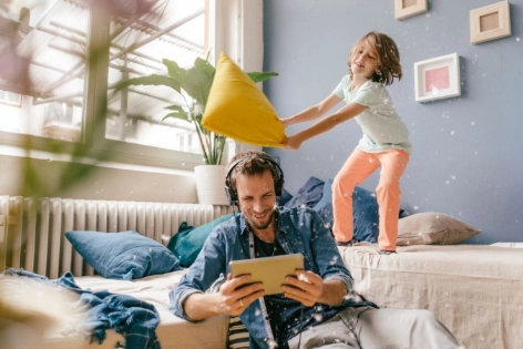 Vater hört Music auf dem Tablet und Kind tobt auf dem Sofa und spielt Kissenschlacht.
