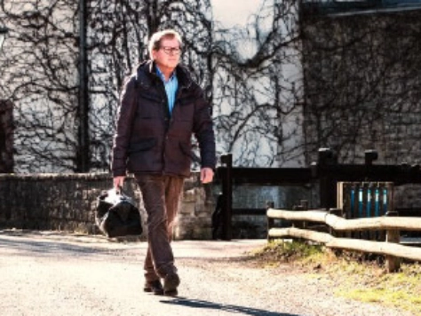 Mann in brauner Jacke und Brille geht auf Kiesweg, kahle Bäume und Holzstapel im Hintergrund