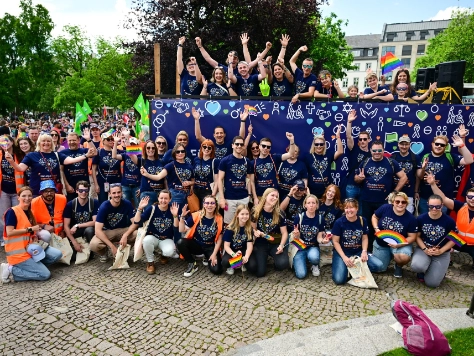 Große Gruppe Menschen in dunkelblauen Shirts posiert jubelnd auf Pflastersteinen vor Gebäuden, viele mit erhobenen Armen.