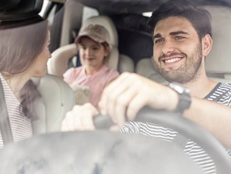 Zwei Personen sitzen lächelnd im Auto, der Fahrer trägt ein gestreiftes Shirt, der Beifahrer eine rosa Kappe und helle Kleidung.