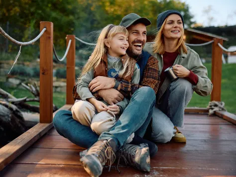 Mutter, Vater und Tochter sitzen auf einem Bootssteg und beschäftigen sich mit der Frage, was ein Kind bis zum 18. Lebensjahr kostet. 